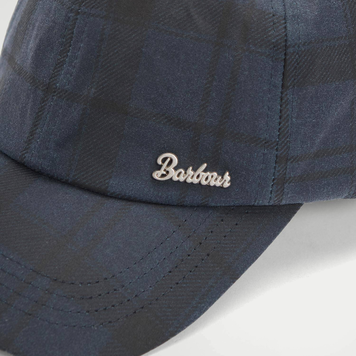 Gros plan de la plaque logo Barbour script en métal et de la visière surpiquée, coton ciré tartan bleu marine