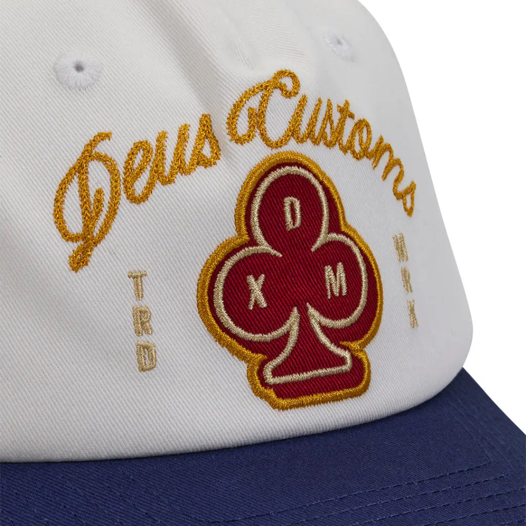 Détail de la broderie point de chaînette dorée Deus Customs et écusson trèfle rouge sur la calotte écrue