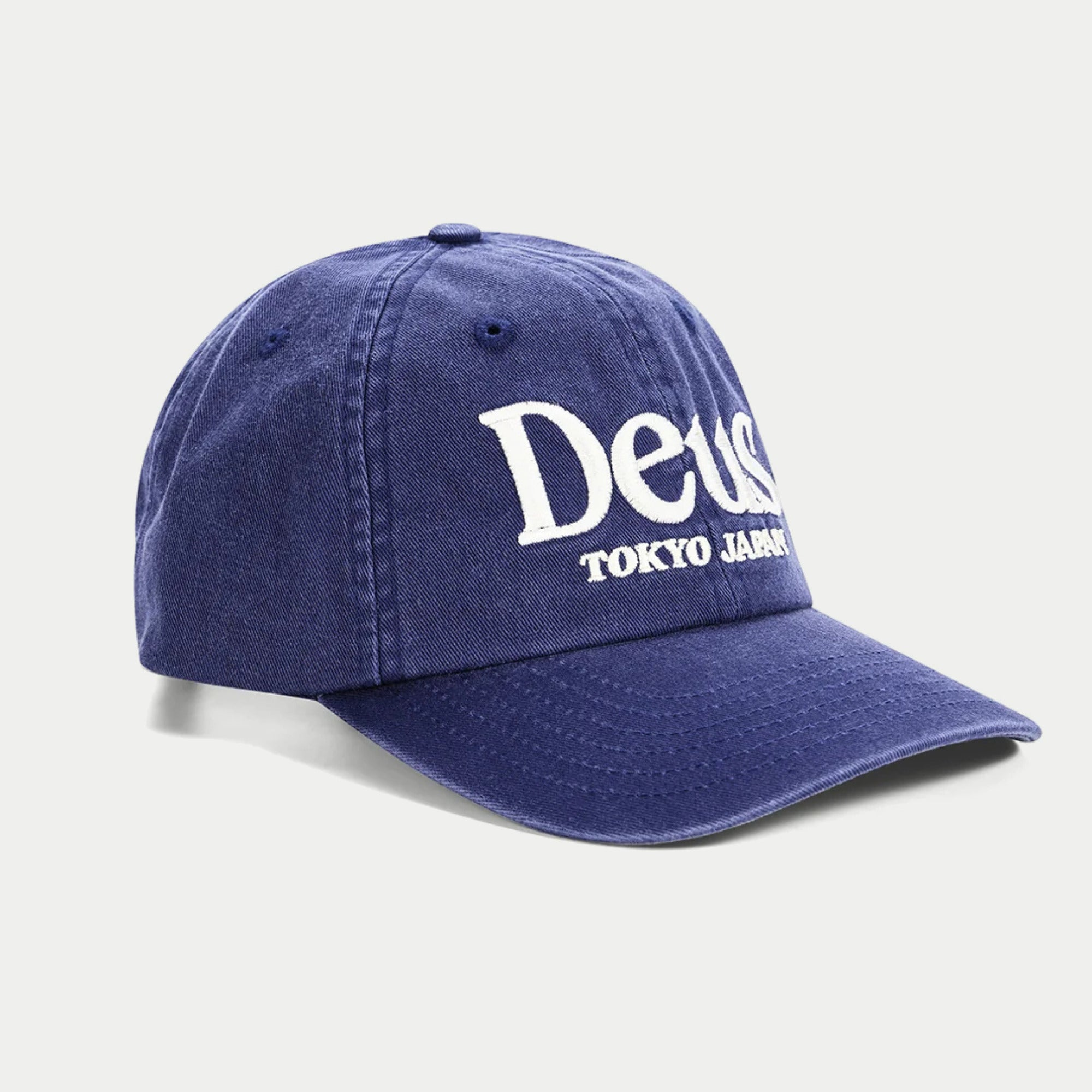 Casquette Metro indigo Deus Ex Machina de trois quarts avant, lettrage sérif Deus et TOKYO JAPAN brodés blanc sur le devant