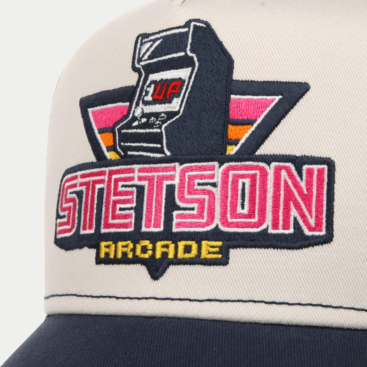 Gros plan de la broderie borne d'arcade 1UP et du lettrage Stetson Arcade rose et jaune sur le devant beige