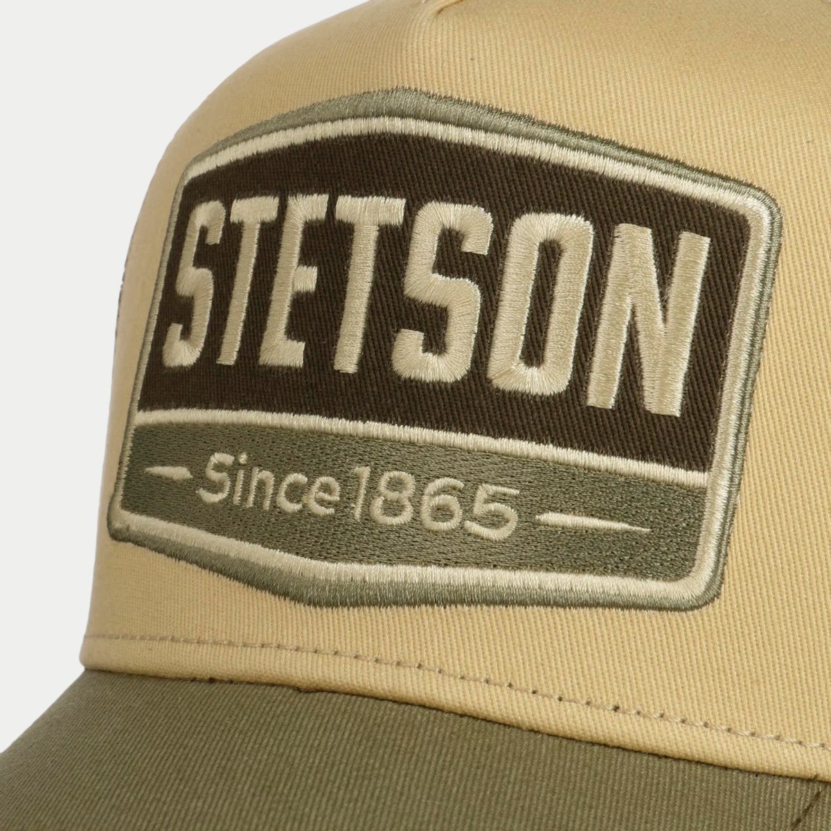 Gros plan sur l'écusson Stetson Since 1865 brodé brun et olive de la casquette trucker Highway, devant beige sable