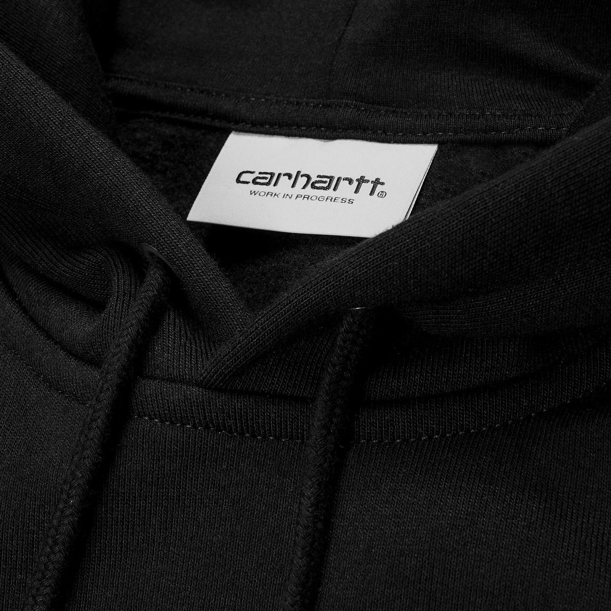Macro de l'étiquette tissée Carhartt Work In Progress dans la capuche noire du Hoodie Chase, cordon et œillet