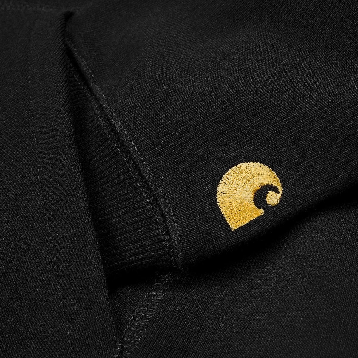 Macro du logo C brodé jaune doré sur la manche noire du Hoodie Chase, juste au-dessus du poignet côtelé