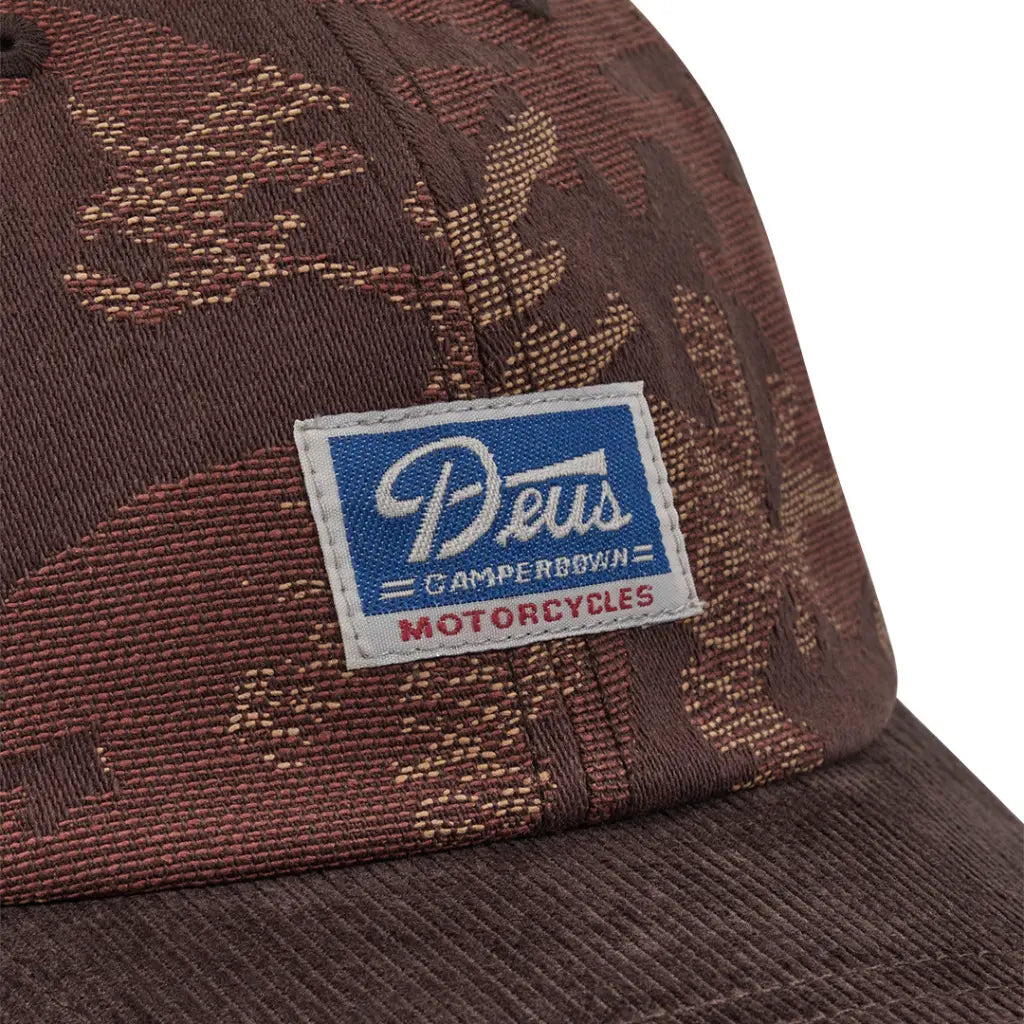 Gros plan du patch tissé bleu Deus Motorcycles sur le jacquard camouflage brun de l'Idaho Cap