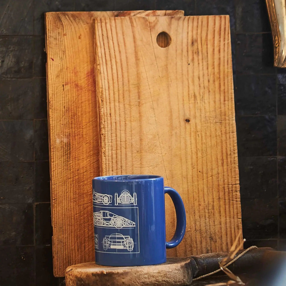 Mug bleu 24h Le Mans posé sur un billot devant des planches en bois, motif blueprint visible