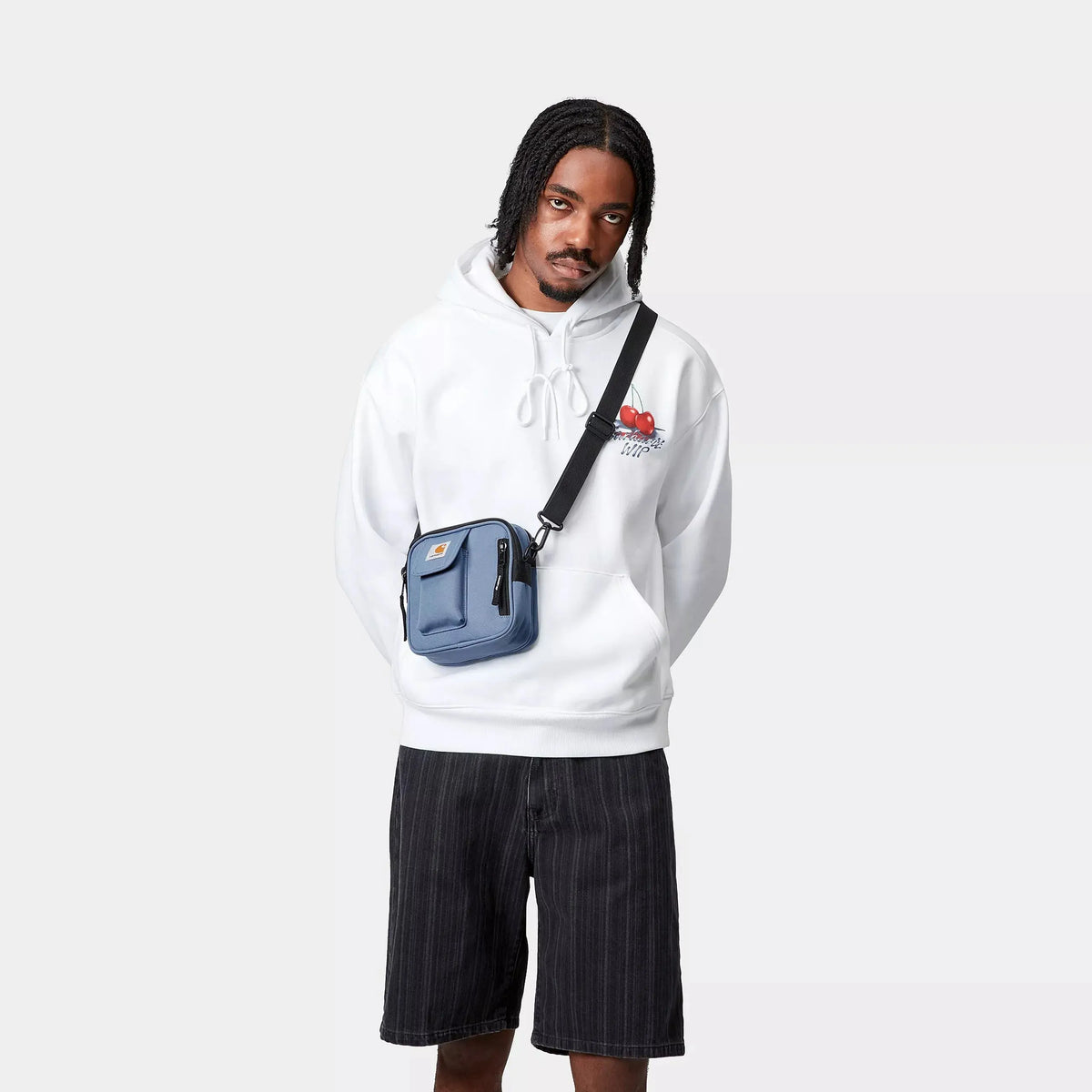 Sacoche Essentials Bag Sorrent Carhartt WIP bleu ardoise portée en bandoulière sur un sweat à capuche blanc