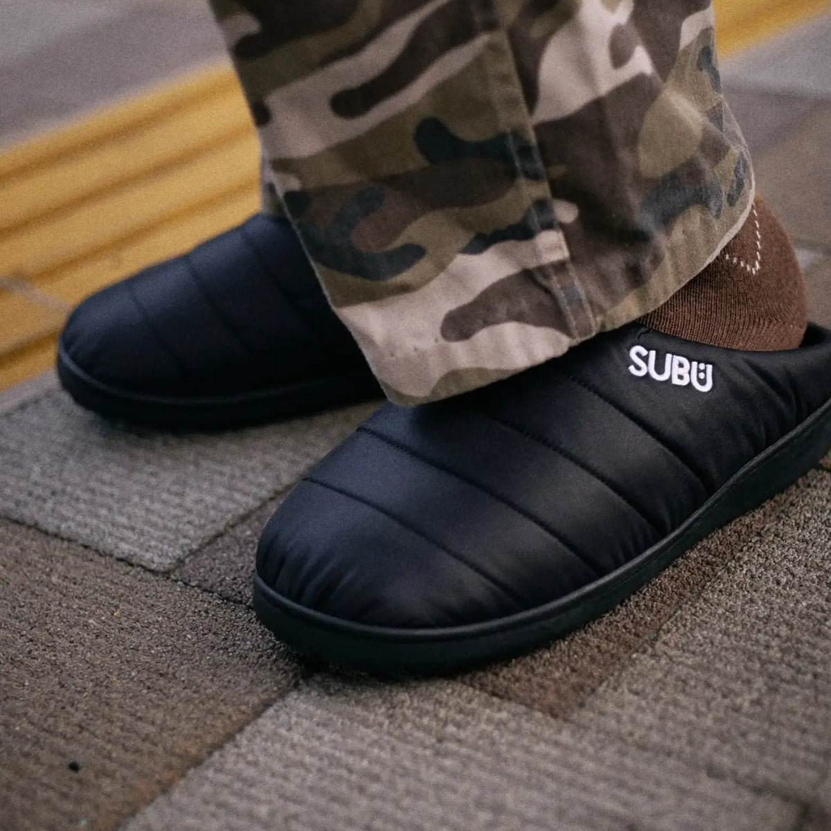 Chaussons noirs portés avec un pantalon camouflage et des chaussettes marron sur un sol carrelé