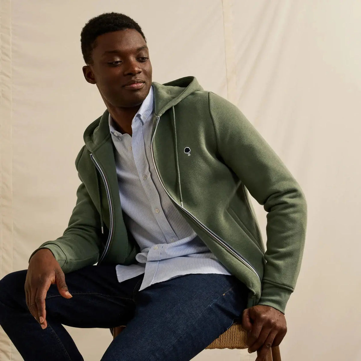 Sweat zippé à capuche kaki porté ouvert sur une chemise rayée bleu clair et un jean brut, capuche à plat