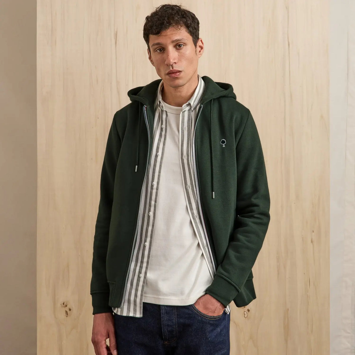 Sweat zippé vert foncé porté ouvert sur une chemise rayée et un t-shirt blanc, cordons de capuche pendants
