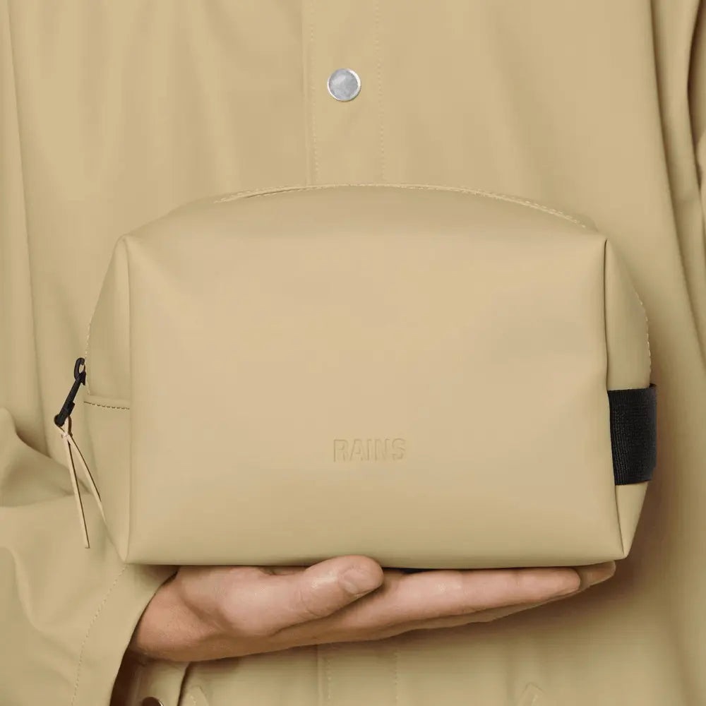 Wash Bag Small Sand tenue en main devant une veste beige sable, patte noire bien visible