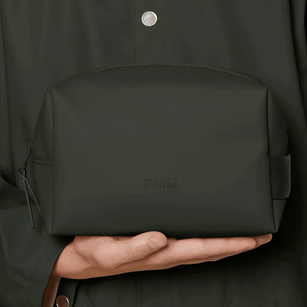 Wash Bag Small vert kaki tenue en main devant une veste vert foncé, format compact